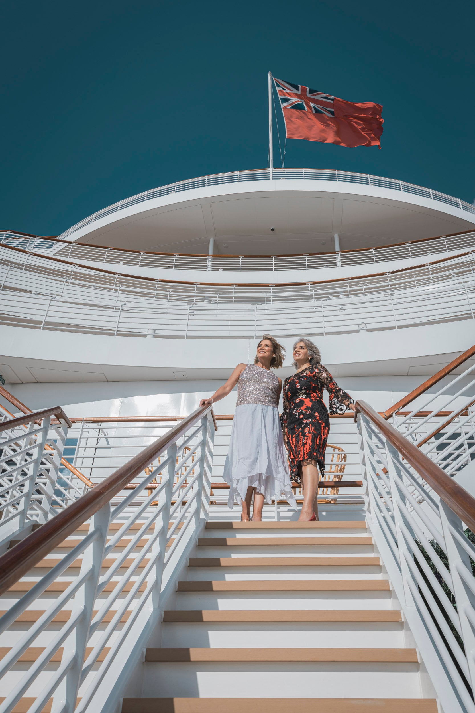 Saga cruise lifestyle photography — two women in cocktail dresses on an exterior deck staircase under the Red Ensign