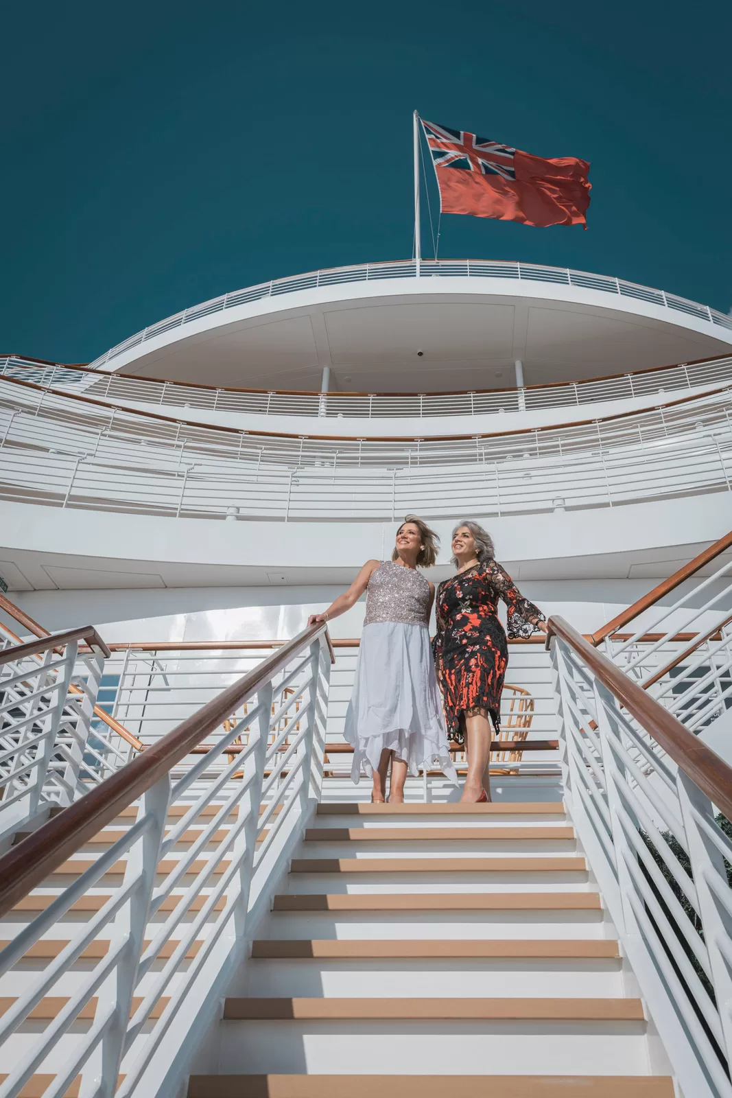 Saga cruise lifestyle photography — two women in cocktail dresses on an exterior deck staircase under the Red Ensign