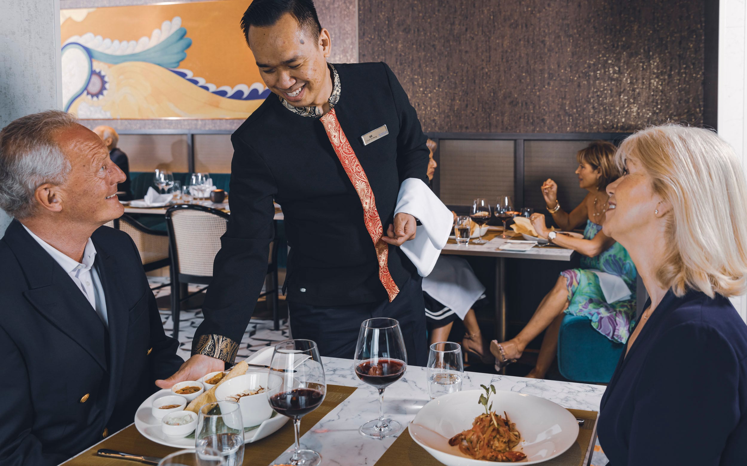 Saga cruise fine-dining photography — waiter serving a pasta course to a smiling couple at their table