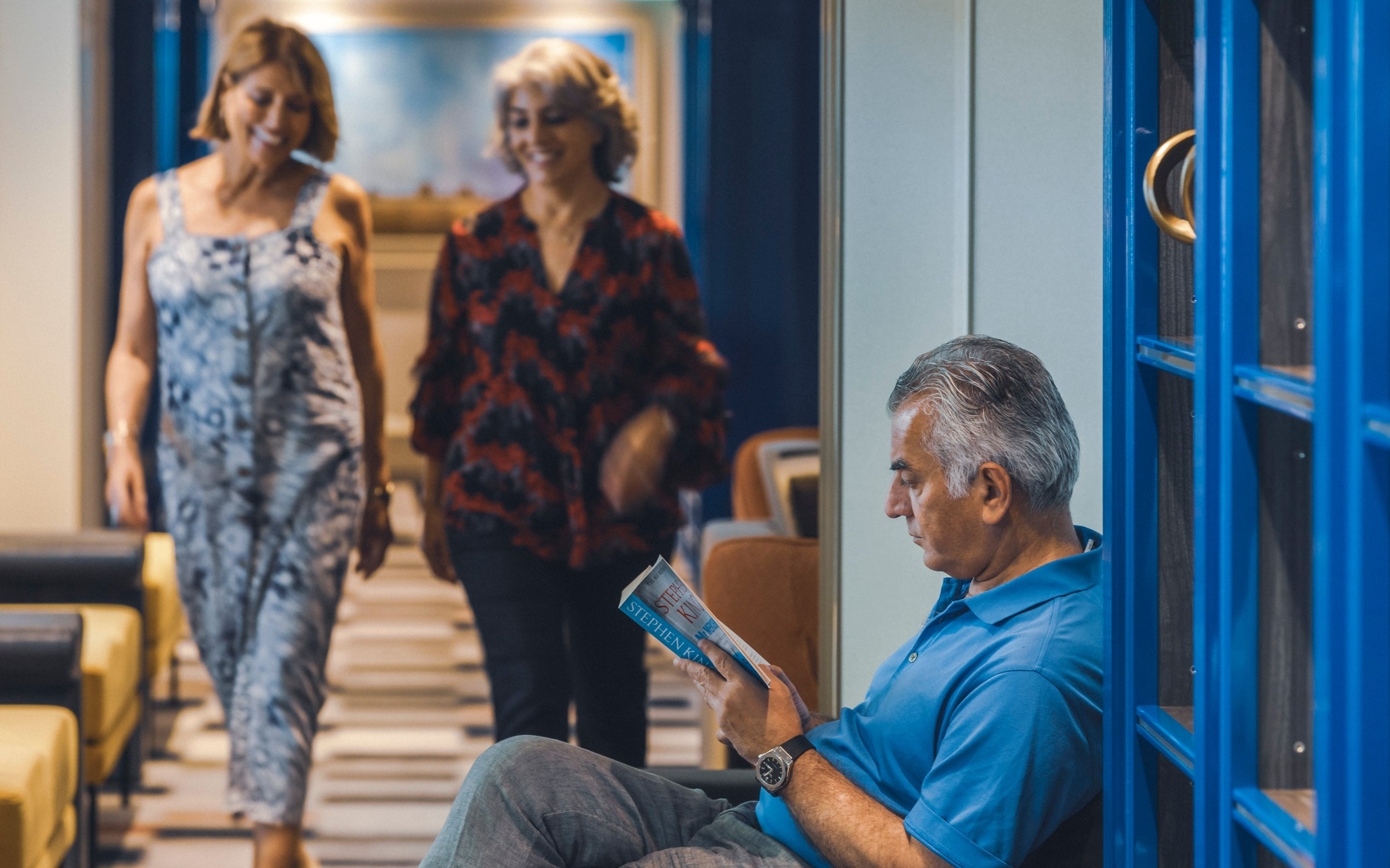 Saga cruise lifestyle photography — man reading a newspaper as two women chat in a ship corridor