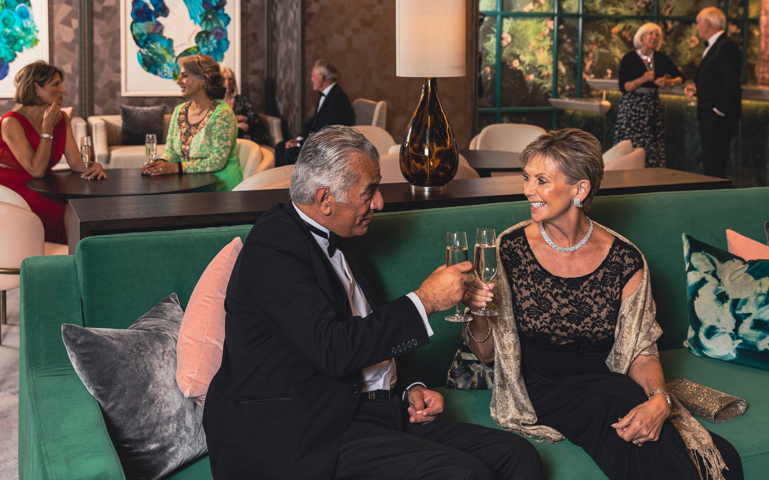 Saga cruise evening photography — formally-dressed couple toasting champagne on a green velvet sofa in a lounge