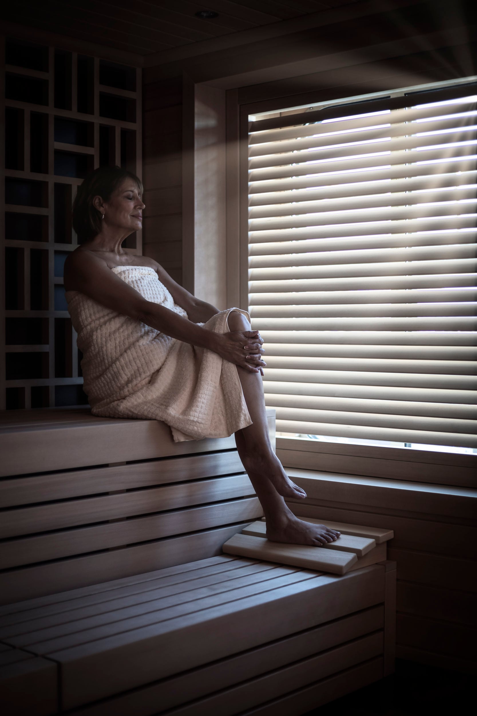Saga cruise spa photography — woman relaxing on a wooden bench inside a sauna, side light through slatted blinds