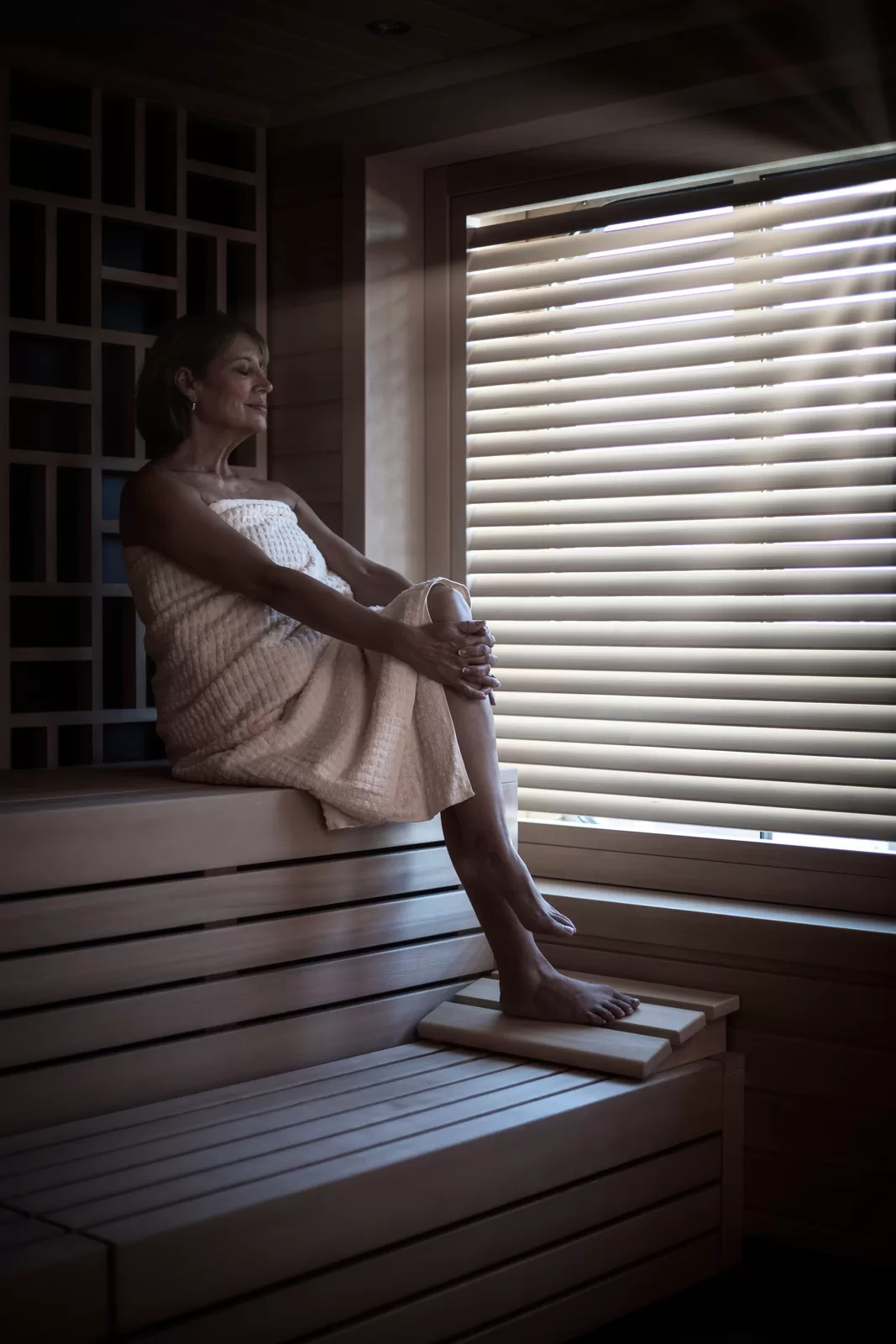 Saga cruise spa photography — woman relaxing on a wooden bench inside a sauna, side light through slatted blinds