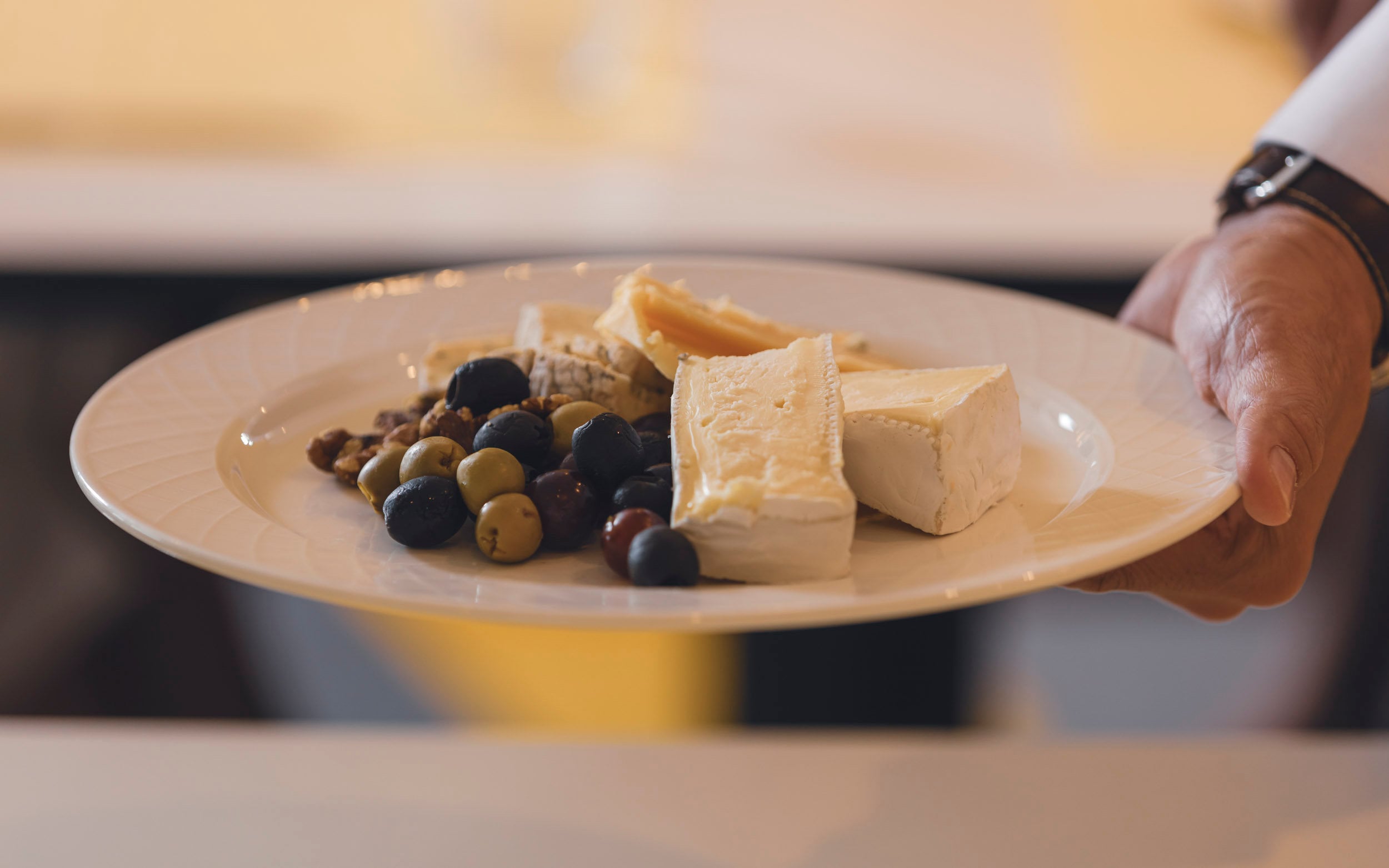 Saga cruise fine-dining photography — cheese-board plate with brie, olives and walnuts held by a waiter