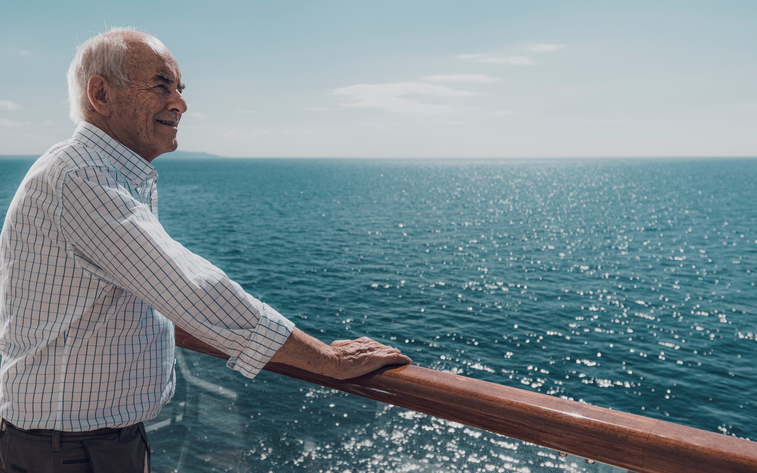 Saga cruise lifestyle photography — older man on the open deck leaning against a polished wooden rail, looking out to sea
