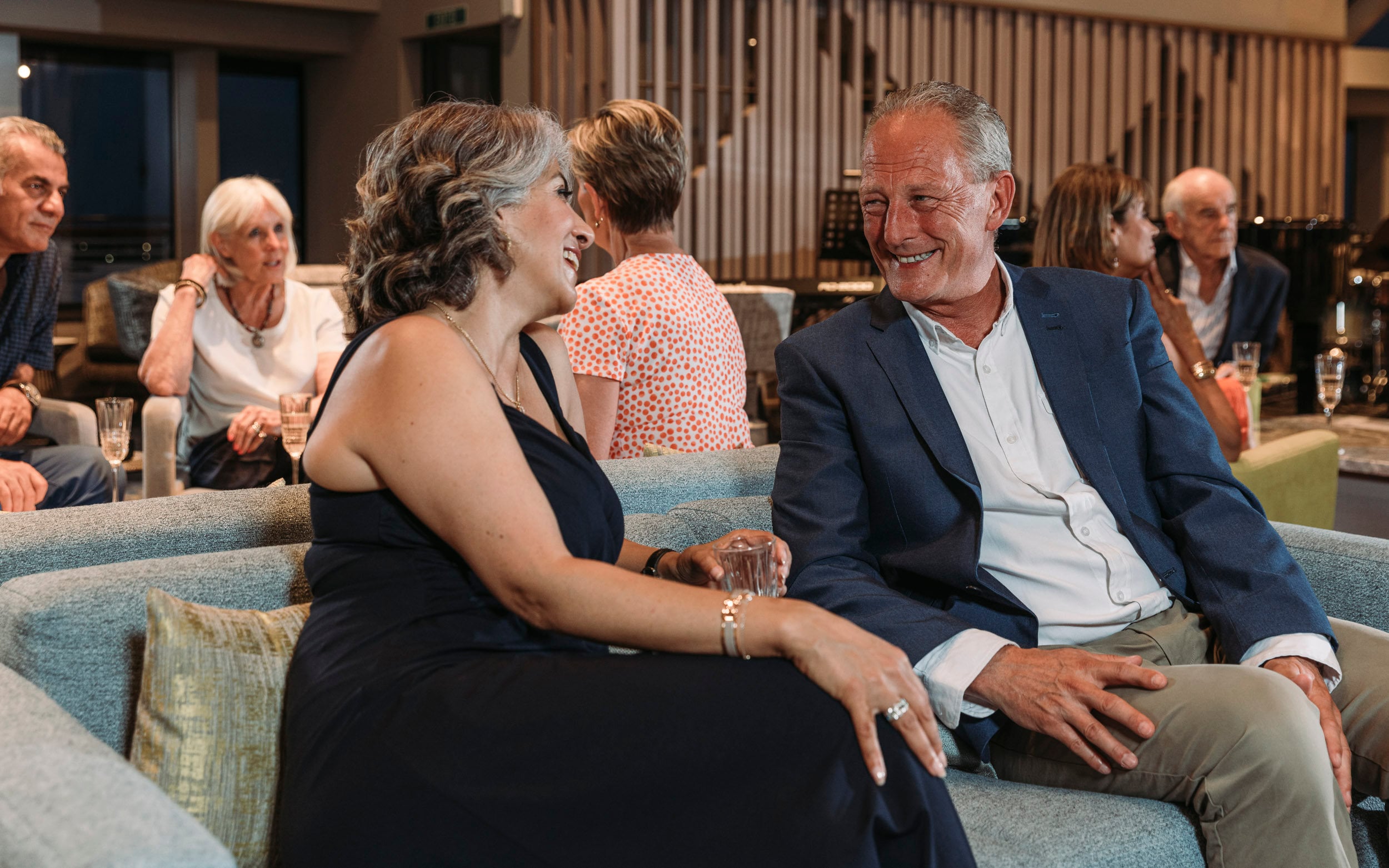 Saga cruise lifestyle photography — couple sharing a laugh on a teal sofa in a busy ship lounge